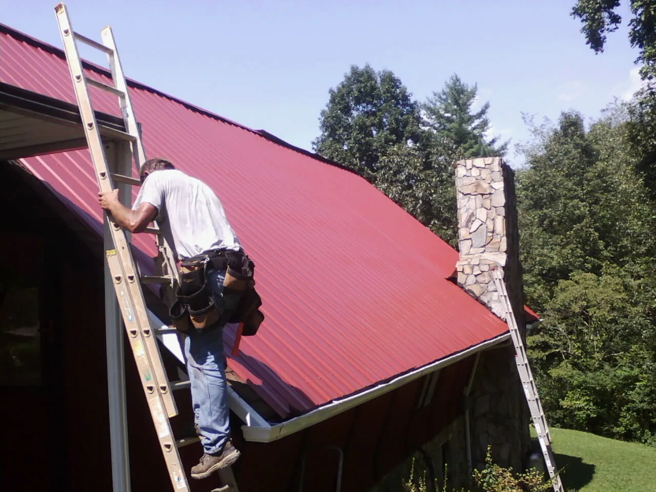 Roofer on a ladder beside a red metal roof and stone chimney — Soffit & Fascia Repair by Roofing Brothers in Donnelly
